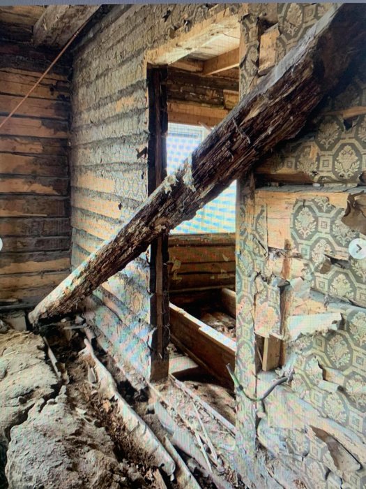 Förfallen interiör av ett gammalt hus med bortfallande timmer och trasigt tapetserade väggar.