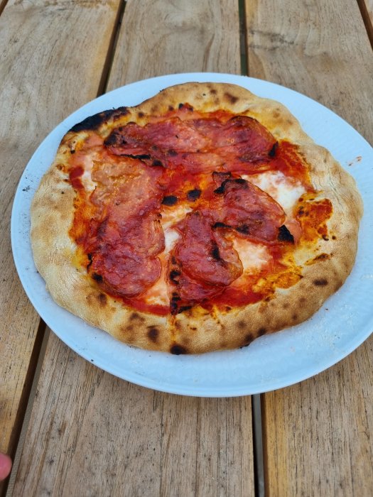 Nybakad pizza med salami på vit tallrik, placerad på ett träbord utomhus.