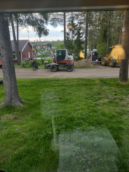 Vy genom fönster på byggplats med grävmaskin och lastbil, med skog och hus i bakgrunden.