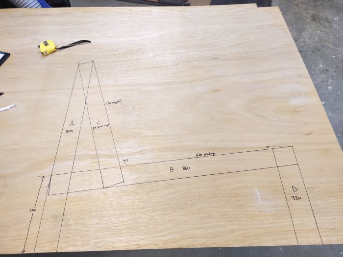 En plywoodskiva med påritade mått och skisser för bygge av soffdelar, bredvid en tumstock.