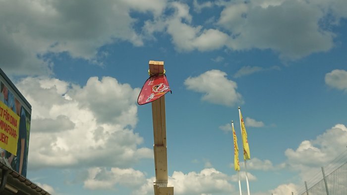 Lutande träregel med band och planka i toppen som håller en röd flagga mot himlen.