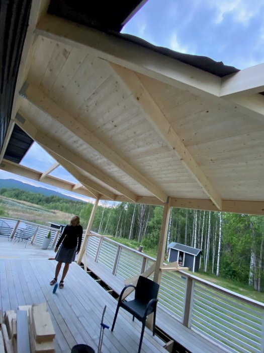 Konstruktion av en inglasad altan med naturligt trä mot en skogsbakgrund, svarta detaljer, och en person som överväger designval.