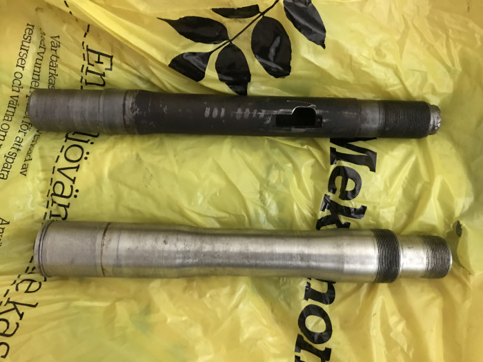 Två metallstyrstammar med gängning ligger på en gul plastpåse, en är nyligen svarvad medan den andra är använd.
