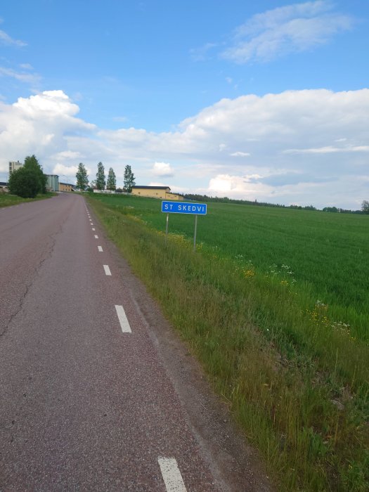 Väg med cykelbana och skylt som visar St Skedvi under en blå himmel med moln.