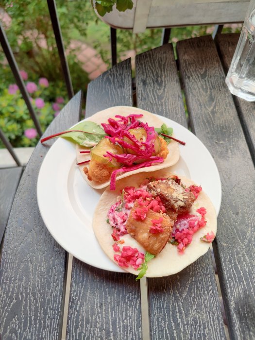 Två tacos med rödlökslag, kött, sallad och tortillabröd på ett vitt fat mot mörkt träbord.