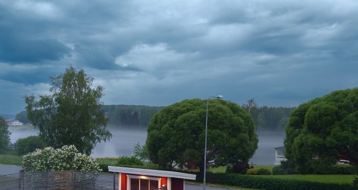 Mörka regnmoln över landskap med dimma över sjö, träd i förgrunden, och hustak.