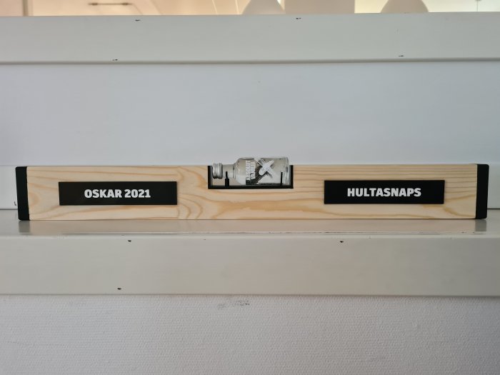 Trähylsa med texten "OSKAR 2021" och "HULTASNAPS", innehållandes en flaska.