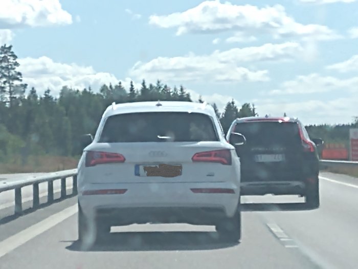 Två bilar kör på en motorväg, en vit Audi framför och svart bil bakom, suddig bildkvalitet.