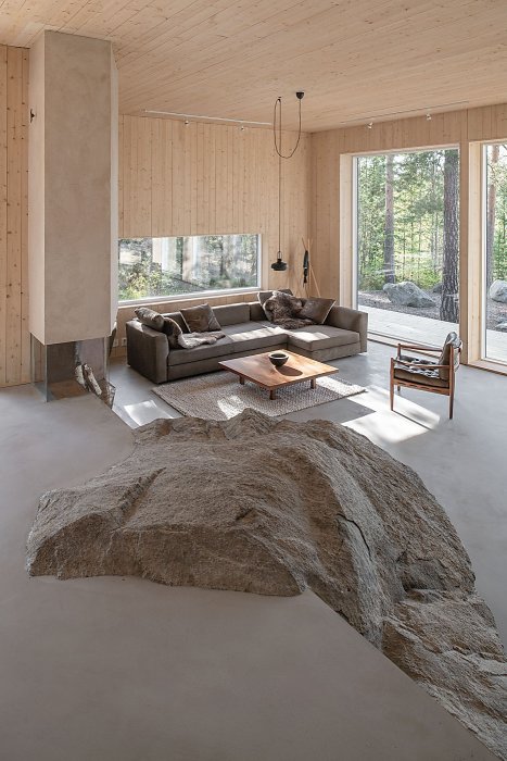 Modernt vardagsrum med naturligt klippblock integrerat i inredningen, träsäten och stora fönster.