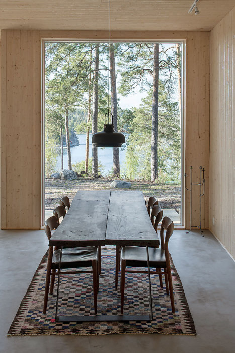Rustikt matbord och stolar i ett ljust rum med stora fönster och utsikt över skog och sjö.
