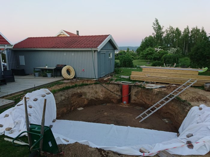 Ett nyligen grävt hål med en plastdräneringsbrunn i centrum, omgivet av byggmaterial, mot en bakgrund av en trädgård och hus.