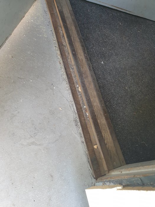 Gammal tröskel med småsten och grus vid en dörröppning, med en slitstark matta och betongtrappan syns.