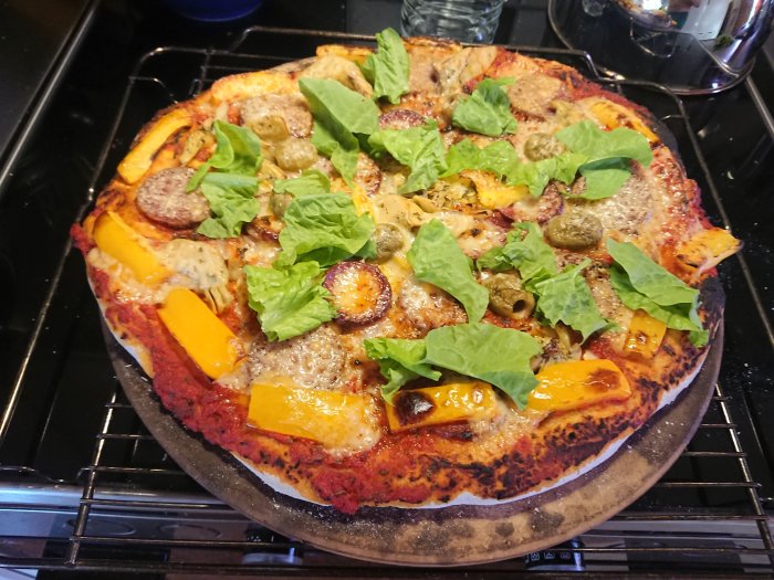 Hembakt pizza med salami, hästkorv, paprika, oliver och sallad på stenplatta i ugn.