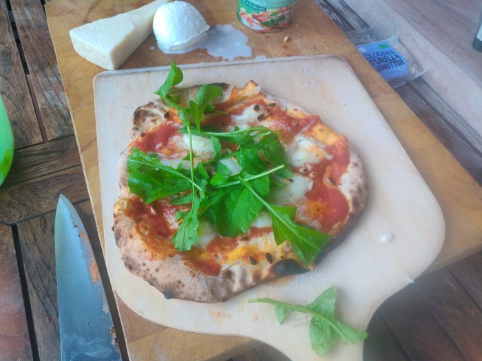 Hembakad pizza med pecorino, pinjenötter och ruccola på skärbräda, kniv och ostar i bakgrunden.