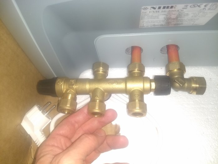 En hand håller en termostatventil framför en värmepanna, vänter vänt mot kameran.