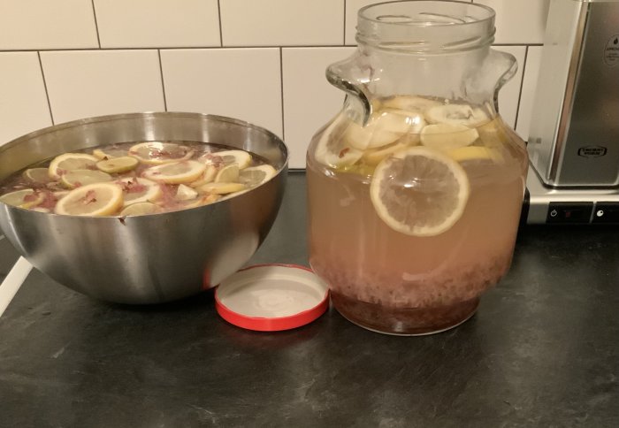Tillredning av syrensaft med citronskivor i en stor metallskål och en glaskanna på ett köksbord.