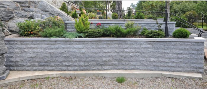 Granitmur av mursten som stödmur i en trädgård med växtlighet ovanpå och staket bakom.