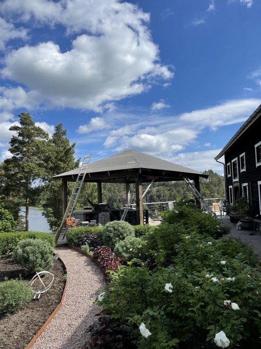 Välansad trädgård med blomsterbäddar framför ett paviljongtak nära vatten, med en stege lutad mot konstruktionen.