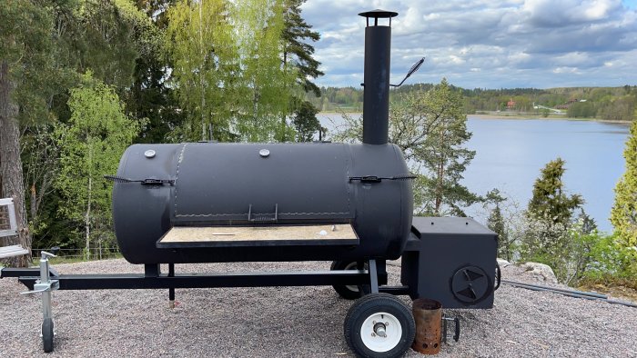 Hembyggd smoker på hjul framför sjöutsikt med skogsmiljö i bakgrunden.