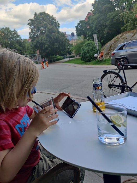 Barn som håller i ett glas vid ett utebord med en flaska tonic och ett tomt glas med is och citronskiva, i en stadsmiljö.