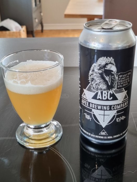 Ölglas fyllt med ljus öl bredvid Apex Brewing Company burk på bord.