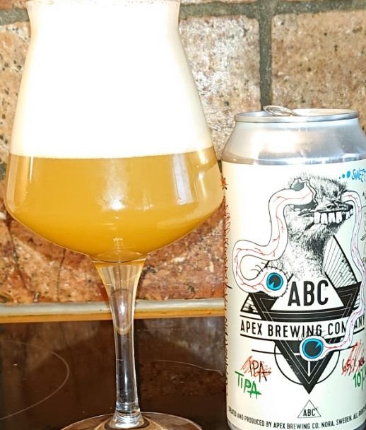 Ett glas fyllt med öl bredvid en Apex Brewing burk med konstnärlig etikett mot en tegelvägg.