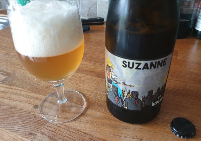 Ölflaska med etiketten "SUZANNE" och ett glas med öl och tjockt skum på ett bord.