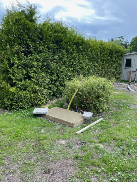 Betongplatta med två stenvikter och en gul vattenslang på en trädgårdstomt i byggfasen.