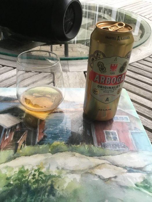 Ölburk av Arboga med text "Original Ale" och en delvis fylld ölglas bredvid på ett bord med bordsduk med landskapsmålning.