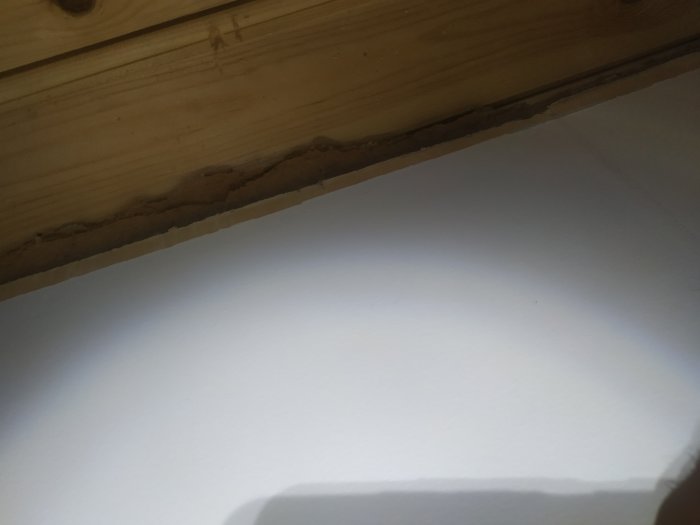 Kanten där en masonit- eller träfiberskiva i taket möter väggen, med en synlig spik och en överlappning.