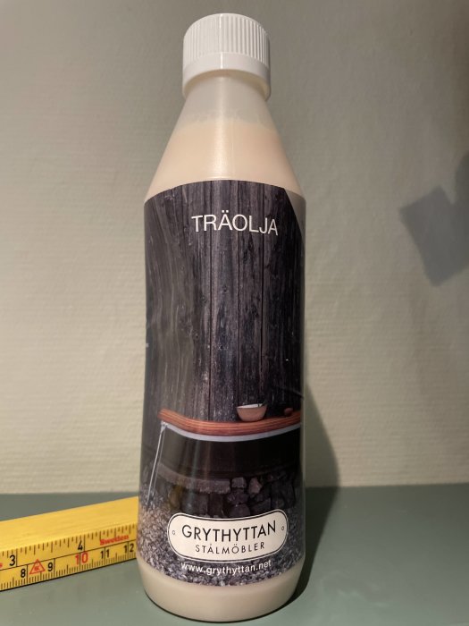 Flaska med Grythyttans original träolja bredvid en tumstock på en bordsyta.