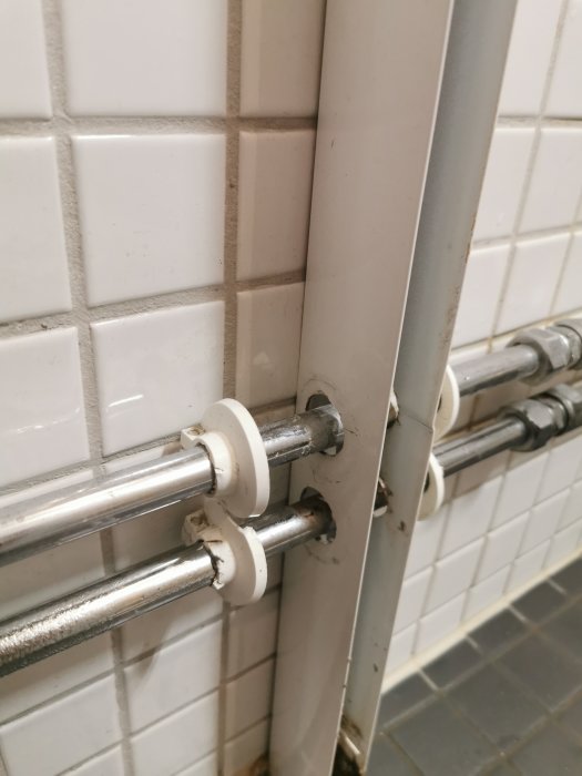 Fästen för duschväggar monterade på vita kakelväggar och rörledningar i ett badrum.