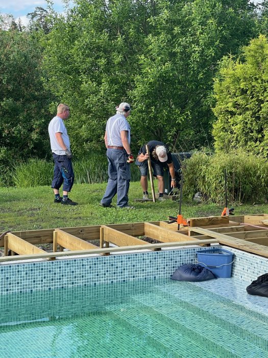 Män arbetar på en halvbyggd träram runt en kaklad pool med grönska i bakgrunden.