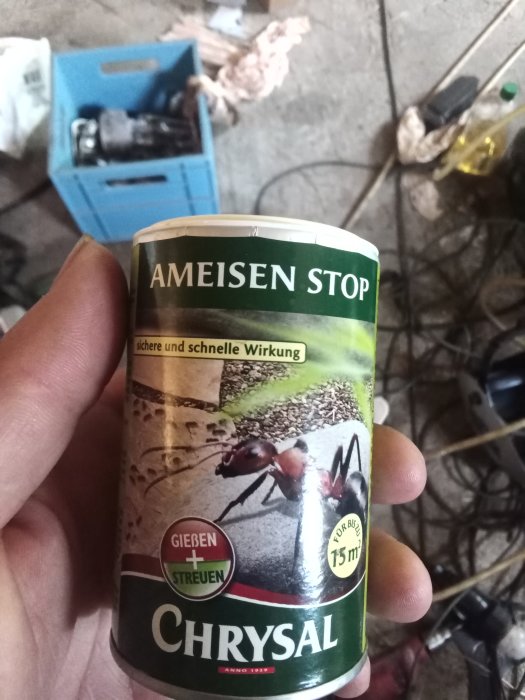 Hand håller en burk med myrbekämpningsmedel "Ameisen Stop" i ett rörigt garage.