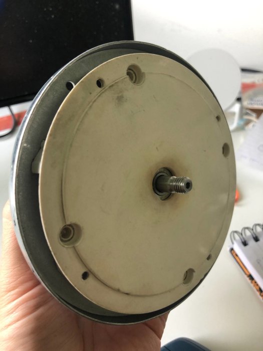 En hand håller en vit plasttallrik med centralt axel och borttagna skruvar, troligen del av en motorrotor.