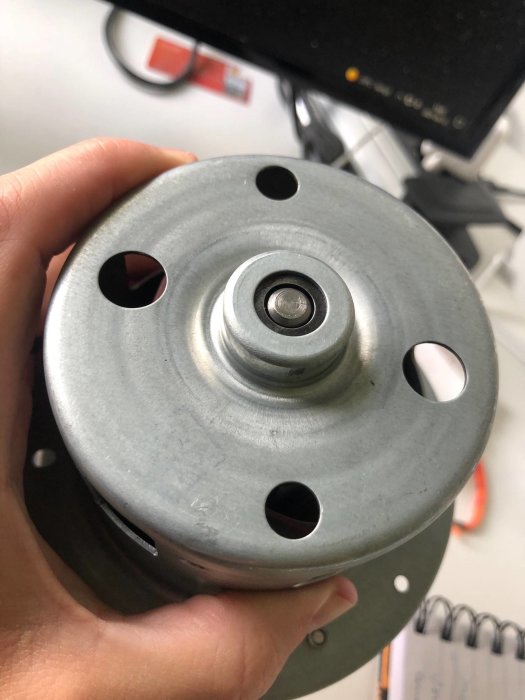 En hand håller en grå plasttallrik med en central axel och borttagna skruvar, del av en rotor.