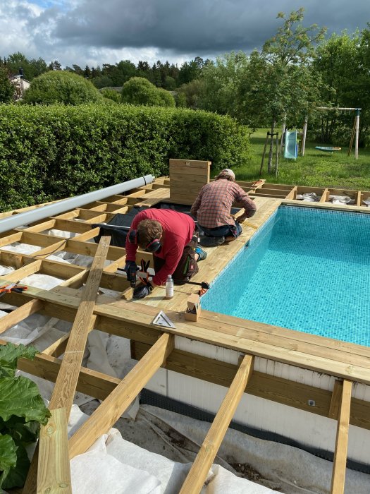 Två personer bygger trädäck kring en fylld swimmingpool i en trädgård.