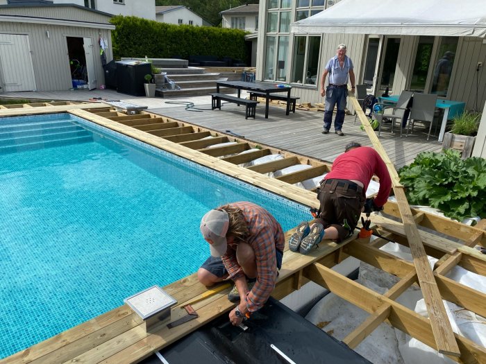 Arbetare som färdigställer trall runt en pool, med en person som övervakar i bakgrunden.