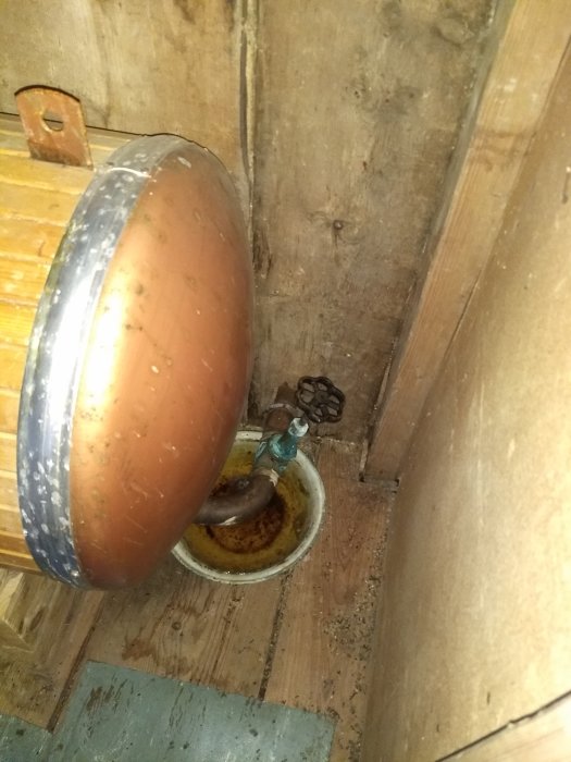 Gammal rostig kran över en gulnad balja som samlar vattendroppar på ett trägolv.