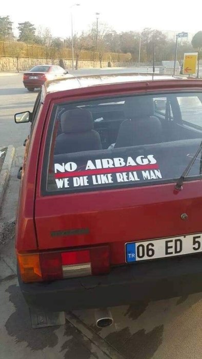 Röd bil parkerad med klistermärke på bakrutan som läser "NO AIRBAGS WE DIE LIKE REAL MAN".