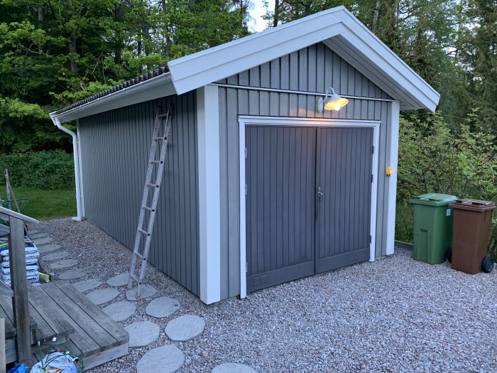 Välmålad garagebyggnad med grå fasad och vit takkant, belyst av en utomhuslampa, med steppstenar och sophinkar framför.