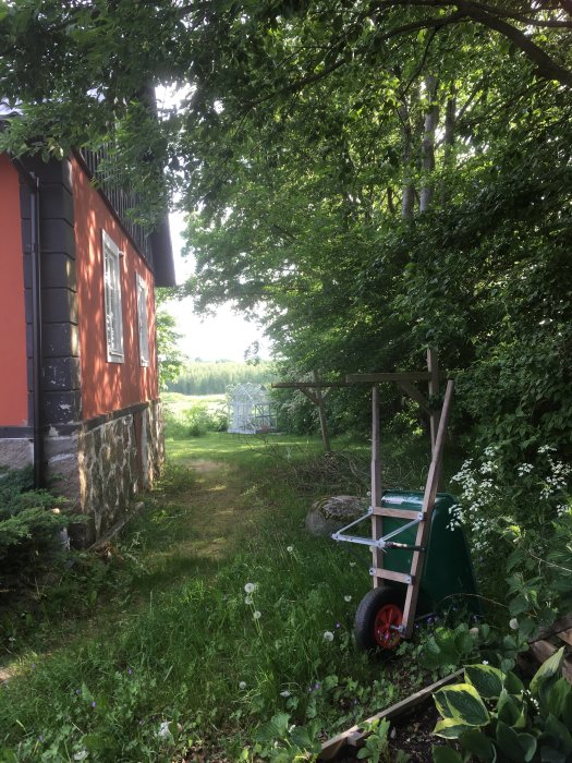 Trädgårdsvagn nära en lummig grusgång och stenläggning med överväxt ogräs vid röd stuga.