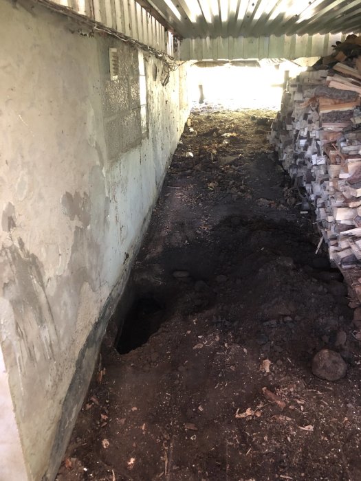 Grund av äldre hus med synlig jord, sten och grävd grop längs husväggen under utstakad plats.