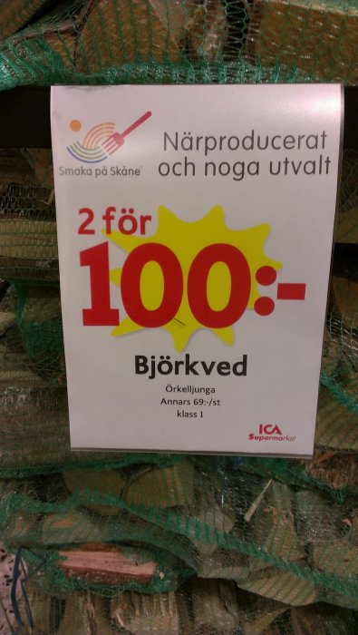 Reklamskylt med erbjudande "2 för 100:- Björkved" framför nätsäckar med ved.