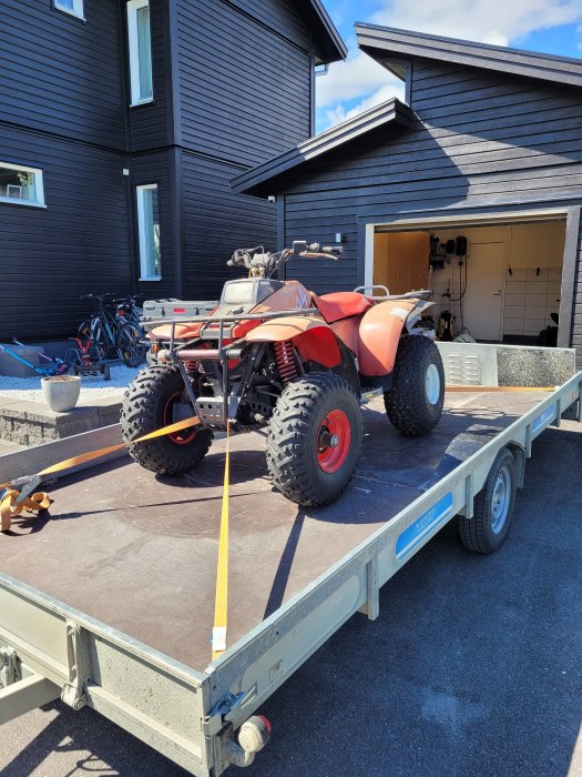 Polaris Trail Boss 250 fyrhjuling på släpvagn framför svart trähus, säkrad med spännband.