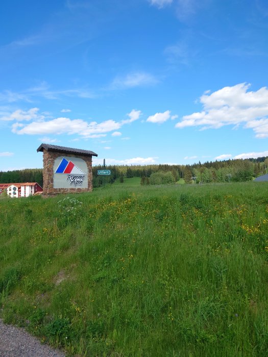 Skylt med företagslogotyp vid en blommande äng med skog i bakgrunden under blå himmel.