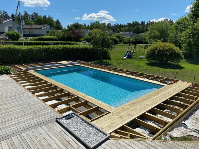 Trädgård med delvis färdigställd trädäck runt en inbyggd pool under solig himmel.