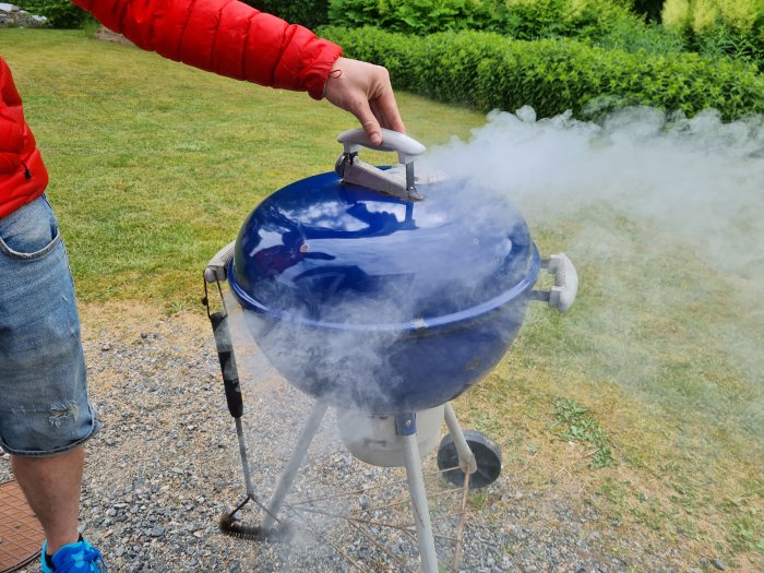 En person lyfter locket på en rykande grill utomhus.