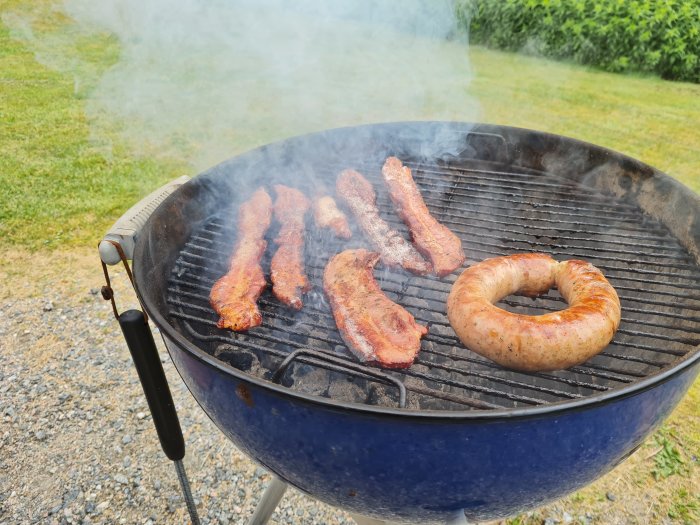Bacon och en Salsiccia-korv grillas på en kolgrill med rök som stiger upp.
