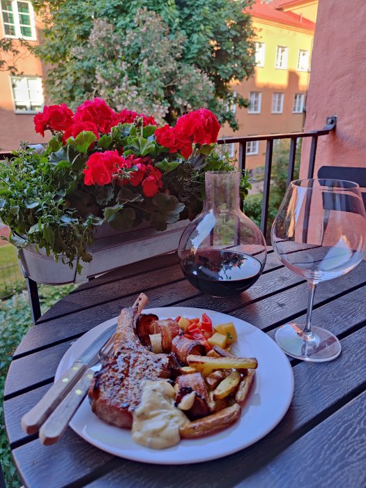 Middagsdukning på balkong med grillad kötträtt, potatis, rödvin och röda blommor i bakgrunden.